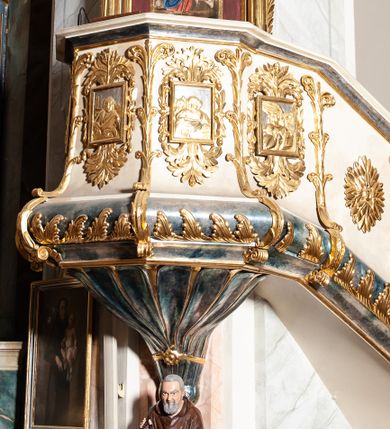 Zdjęcie nr 1: Ambona przyścienna, wisząca ustawiona przy zachodnim filarze łuku tęczowego; dostępna jednobiegowymi schodami z balustradą od strony prezbiterium. Kosz na planie ośmioboku, na puklowanej podstawie spiętej u dołu złoconą obręczą z kwiatem. U dołu wydzielony silnie występującym ćwierćwałkiem z nałożoną złoconą dekoracją w formie liści akantu;  u góry zamknięty profilowanym gzymsem.  Ścianki artykułowane łodygą roślinną o pionowym przebiegu. W polach prostokątne płyciny mieszczące płaskorzeźbione portrety czterech ewangelistów wraz z ich atrybutami umieszczone na tle symetrycznego  ornamentu roślinnego. Balustrada schodów pełna; pola flankowane analogiczną jak na koszu łodygą, pośrodku pól dwie wielolistne rozety. Zaplecek flankowany jońskimi kolumnami o złoconych, kanelowanych trzonach wspierających baldachim oraz uszami utworzonymi z rocaille&#039;ów, kratki regencyjnej oraz liści laurowych. W polu prostokątna płycina zamknięta łukiem wklęsło-wypukłym z uskokiem mieszcząca obraz Dobrego Pasterza malowany olejem na desce. Baldachim ośmioboczny, profilowany z podwieszonym u dołu lambrekinem. W partii fryzu girlandy podwieszone na kokardach z wstęgi regencyjnej. W zwieńczeniu ażurowa  struktura utworzona z wolut wspierająca postument z pełnoplastyczną, polichromowaną figurą św. Pawła.
Polichromia jasnobeżowa, miejscami marmoryzowana w odcieniach morskiego błękitu; ornamentyka oraz detale architektoniczne złocone i srebrzone.