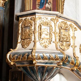 Zdjęcie nr 1: Ambona przyścienna, wisząca ustawiona przy zachodnim filarze łuku tęczowego; dostępna jednobiegowymi schodami z balustradą od strony prezbiterium. Kosz na planie ośmioboku, na puklowanej podstawie spiętej u dołu złoconą obręczą z kwiatem. U dołu wydzielony silnie występującym ćwierćwałkiem z nałożoną złoconą dekoracją w formie liści akantu;  u góry zamknięty profilowanym gzymsem.  Ścianki artykułowane łodygą roślinną o pionowym przebiegu. W polach prostokątne płyciny mieszczące płaskorzeźbione portrety czterech ewangelistów wraz z ich atrybutami umieszczone na tle symetrycznego  ornamentu roślinnego. Balustrada schodów pełna; pola flankowane analogiczną jak na koszu łodygą, pośrodku pól dwie wielolistne rozety. Zaplecek flankowany jońskimi kolumnami o złoconych, kanelowanych trzonach wspierających baldachim oraz uszami utworzonymi z rocaille&#039;ów, kratki regencyjnej oraz liści laurowych. W polu prostokątna płycina zamknięta łukiem wklęsło-wypukłym z uskokiem mieszcząca obraz Dobrego Pasterza malowany olejem na desce. Baldachim ośmioboczny, profilowany z podwieszonym u dołu lambrekinem. W partii fryzu girlandy podwieszone na kokardach z wstęgi regencyjnej. W zwieńczeniu ażurowa  struktura utworzona z wolut wspierająca postument z pełnoplastyczną, polichromowaną figurą św. Pawła.
Polichromia jasnobeżowa, miejscami marmoryzowana w odcieniach morskiego błękitu; ornamentyka oraz detale architektoniczne złocone i srebrzone.