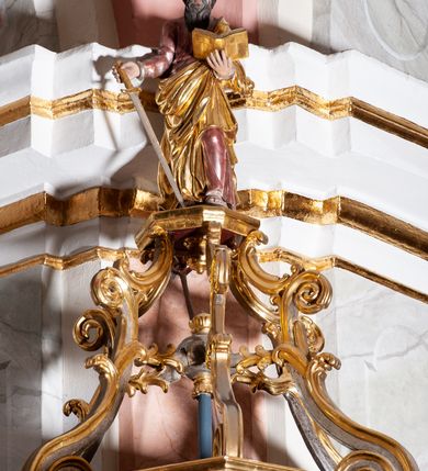 Zdjęcie nr 7: Ambona przyścienna, wisząca ustawiona przy zachodnim filarze łuku tęczowego; dostępna jednobiegowymi schodami z balustradą od strony prezbiterium. Kosz na planie ośmioboku, na puklowanej podstawie spiętej u dołu złoconą obręczą z kwiatem. U dołu wydzielony silnie występującym ćwierćwałkiem z nałożoną złoconą dekoracją w formie liści akantu;  u góry zamknięty profilowanym gzymsem.  Ścianki artykułowane łodygą roślinną o pionowym przebiegu. W polach prostokątne płyciny mieszczące płaskorzeźbione portrety czterech ewangelistów wraz z ich atrybutami umieszczone na tle symetrycznego  ornamentu roślinnego. Balustrada schodów pełna; pola flankowane analogiczną jak na koszu łodygą, pośrodku pól dwie wielolistne rozety. Zaplecek flankowany jońskimi kolumnami o złoconych, kanelowanych trzonach wspierających baldachim oraz uszami utworzonymi z rocaille'ów, kratki regencyjnej oraz liści laurowych. W polu prostokątna płycina zamknięta łukiem wklęsło-wypukłym z uskokiem mieszcząca obraz Dobrego Pasterza malowany olejem na desce. Baldachim ośmioboczny, profilowany z podwieszonym u dołu lambrekinem. W partii fryzu girlandy podwieszone na kokardach z wstęgi regencyjnej. W zwieńczeniu ażurowa  struktura utworzona z wolut wspierająca postument z pełnoplastyczną, polichromowaną figurą św. Pawła.
Polichromia jasnobeżowa, miejscami marmoryzowana w odcieniach morskiego błękitu; ornamentyka oraz detale architektoniczne złocone i srebrzone.