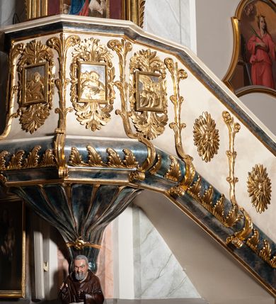 Zdjęcie nr 4: Ambona przyścienna, wisząca ustawiona przy zachodnim filarze łuku tęczowego; dostępna jednobiegowymi schodami z balustradą od strony prezbiterium. Kosz na planie ośmioboku, na puklowanej podstawie spiętej u dołu złoconą obręczą z kwiatem. U dołu wydzielony silnie występującym ćwierćwałkiem z nałożoną złoconą dekoracją w formie liści akantu;  u góry zamknięty profilowanym gzymsem.  Ścianki artykułowane łodygą roślinną o pionowym przebiegu. W polach prostokątne płyciny mieszczące płaskorzeźbione portrety czterech ewangelistów wraz z ich atrybutami umieszczone na tle symetrycznego  ornamentu roślinnego. Balustrada schodów pełna; pola flankowane analogiczną jak na koszu łodygą, pośrodku pól dwie wielolistne rozety. Zaplecek flankowany jońskimi kolumnami o złoconych, kanelowanych trzonach wspierających baldachim oraz uszami utworzonymi z rocaille&#039;ów, kratki regencyjnej oraz liści laurowych. W polu prostokątna płycina zamknięta łukiem wklęsło-wypukłym z uskokiem mieszcząca obraz Dobrego Pasterza malowany olejem na desce. Baldachim ośmioboczny, profilowany z podwieszonym u dołu lambrekinem. W partii fryzu girlandy podwieszone na kokardach z wstęgi regencyjnej. W zwieńczeniu ażurowa  struktura utworzona z wolut wspierająca postument z pełnoplastyczną, polichromowaną figurą św. Pawła.
Polichromia jasnobeżowa, miejscami marmoryzowana w odcieniach morskiego błękitu; ornamentyka oraz detale architektoniczne złocone i srebrzone.