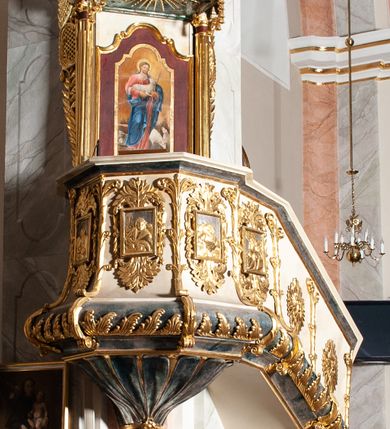 Zdjęcie nr 3: Ambona przyścienna, wisząca ustawiona przy zachodnim filarze łuku tęczowego; dostępna jednobiegowymi schodami z balustradą od strony prezbiterium. Kosz na planie ośmioboku, na puklowanej podstawie spiętej u dołu złoconą obręczą z kwiatem. U dołu wydzielony silnie występującym ćwierćwałkiem z nałożoną złoconą dekoracją w formie liści akantu;  u góry zamknięty profilowanym gzymsem.  Ścianki artykułowane łodygą roślinną o pionowym przebiegu. W polach prostokątne płyciny mieszczące płaskorzeźbione portrety czterech ewangelistów wraz z ich atrybutami umieszczone na tle symetrycznego  ornamentu roślinnego. Balustrada schodów pełna; pola flankowane analogiczną jak na koszu łodygą, pośrodku pól dwie wielolistne rozety. Zaplecek flankowany jońskimi kolumnami o złoconych, kanelowanych trzonach wspierających baldachim oraz uszami utworzonymi z rocaille'ów, kratki regencyjnej oraz liści laurowych. W polu prostokątna płycina zamknięta łukiem wklęsło-wypukłym z uskokiem mieszcząca obraz Dobrego Pasterza malowany olejem na desce. Baldachim ośmioboczny, profilowany z podwieszonym u dołu lambrekinem. W partii fryzu girlandy podwieszone na kokardach z wstęgi regencyjnej. W zwieńczeniu ażurowa  struktura utworzona z wolut wspierająca postument z pełnoplastyczną, polichromowaną figurą św. Pawła.
Polichromia jasnobeżowa, miejscami marmoryzowana w odcieniach morskiego błękitu; ornamentyka oraz detale architektoniczne złocone i srebrzone.