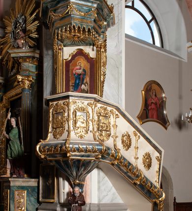 Zdjęcie nr 2: Ambona przyścienna, wisząca ustawiona przy zachodnim filarze łuku tęczowego; dostępna jednobiegowymi schodami z balustradą od strony prezbiterium. Kosz na planie ośmioboku, na puklowanej podstawie spiętej u dołu złoconą obręczą z kwiatem. U dołu wydzielony silnie występującym ćwierćwałkiem z nałożoną złoconą dekoracją w formie liści akantu;  u góry zamknięty profilowanym gzymsem.  Ścianki artykułowane łodygą roślinną o pionowym przebiegu. W polach prostokątne płyciny mieszczące płaskorzeźbione portrety czterech ewangelistów wraz z ich atrybutami umieszczone na tle symetrycznego  ornamentu roślinnego. Balustrada schodów pełna; pola flankowane analogiczną jak na koszu łodygą, pośrodku pól dwie wielolistne rozety. Zaplecek flankowany jońskimi kolumnami o złoconych, kanelowanych trzonach wspierających baldachim oraz uszami utworzonymi z rocaille&#039;ów, kratki regencyjnej oraz liści laurowych. W polu prostokątna płycina zamknięta łukiem wklęsło-wypukłym z uskokiem mieszcząca obraz Dobrego Pasterza malowany olejem na desce. Baldachim ośmioboczny, profilowany z podwieszonym u dołu lambrekinem. W partii fryzu girlandy podwieszone na kokardach z wstęgi regencyjnej. W zwieńczeniu ażurowa  struktura utworzona z wolut wspierająca postument z pełnoplastyczną, polichromowaną figurą św. Pawła.
Polichromia jasnobeżowa, miejscami marmoryzowana w odcieniach morskiego błękitu; ornamentyka oraz detale architektoniczne złocone i srebrzone.