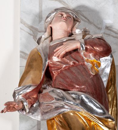 Zdjęcie nr 4: Figura drążona, silnie ścięta z tyłu, przedstawiająca św. Annę. Święta ustawiona frontalnie, w kontrapoście, z prawą nogą ugiętą w kolanie i wspartą na stopniu, o silnie skręconej sylwetce, z lewą ręką złożoną na piersi, prawą odsuniętą w bok. Głowa skierowana w górę, delikatnie przechylona na lewe ramię. Twarz szczupła o wyraźnych rysach, oczy zwrócone ku górze, usta rozchylone. Święta jest ubrana w długą, czerwoną suknię, przepasaną w talii, u spodu obficie marszczoną oraz złocony płaszcz, srebrzony na podbiciu, założony na głowę, opadający na plecy i ramiona, zawinięty na prawej nodze; obficie drapowany. W partiach ciała polichromia naturalistyczna, płaszcz złocony, podbicie srebrzone.