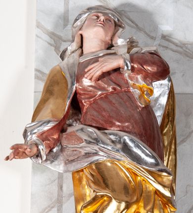 Zdjęcie nr 1: Figura drążona, silnie ścięta z tyłu, przedstawiająca św. Annę. Święta ustawiona frontalnie, w kontrapoście, z prawą nogą ugiętą w kolanie i wspartą na stopniu, o silnie skręconej sylwetce, z lewą ręką złożoną na piersi, prawą odsuniętą w bok. Głowa skierowana w górę, delikatnie przechylona na lewe ramię. Twarz szczupła o wyraźnych rysach, oczy zwrócone ku górze, usta rozchylone. Święta jest ubrana w długą, czerwoną suknię, przepasaną w talii, u spodu obficie marszczoną oraz złocony płaszcz, srebrzony na podbiciu, założony na głowę, opadający na plecy i ramiona, zawinięty na prawej nodze; obficie drapowany. W partiach ciała polichromia naturalistyczna, płaszcz złocony, podbicie srebrzone.