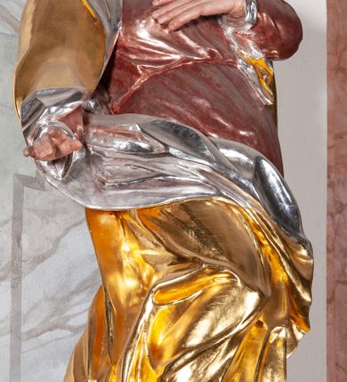 Zdjęcie nr 3: Figura drążona, silnie ścięta z tyłu, przedstawiająca św. Annę. Święta ustawiona frontalnie, w kontrapoście, z prawą nogą ugiętą w kolanie i wspartą na stopniu, o silnie skręconej sylwetce, z lewą ręką złożoną na piersi, prawą odsuniętą w bok. Głowa skierowana w górę, delikatnie przechylona na lewe ramię. Twarz szczupła o wyraźnych rysach, oczy zwrócone ku górze, usta rozchylone. Święta jest ubrana w długą, czerwoną suknię, przepasaną w talii, u spodu obficie marszczoną oraz złocony płaszcz, srebrzony na podbiciu, założony na głowę, opadający na plecy i ramiona, zawinięty na prawej nodze; obficie drapowany. W partiach ciała polichromia naturalistyczna, płaszcz złocony, podbicie srebrzone.