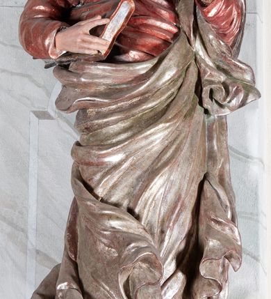 Zdjęcie nr 3: Figura drążona, mocno ścięta z tyłu, przedstawiająca św. Joachima. Święty ustawiony frontalnie, lewa ręka uniesiona i wysunięta przed siebie, a prawą, ugiętą podtrzymuje przy boku księgę. Głowę ma skierowaną w prawą stronę, delikatnie pochyloną. Twarz szeroka z długim nosem o niewielkich oczach patrzących w bok oraz delikatnie rozchylonych ustach; okolona długą brodą w postaci silnie skręconych dwóch pukli opadających diagonalnie na pierś. Włosy długie z przedziałkiem na środku głowy, kręcone, opadają na plecy i lewe ramię. Święty ubrany jest w czerwoną, silnie przylegającą do ciała suknię z wąskimi rękawami oraz brązowy płaszcz przerzucony przez lewe ramię, opadający przez plecy, otaczający postać i odsłaniający klatkę piersiową. Tkanina ekspresyjnie drapowana, obficie fałdowana z rozwianym spodem. Polichromia naturalistyczna w partiach ciała.