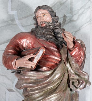 Zdjęcie nr 4: Figura drążona, mocno ścięta z tyłu, przedstawiająca św. Joachima. Święty ustawiony frontalnie, lewa ręka uniesiona i wysunięta przed siebie, a prawą, ugiętą podtrzymuje przy boku księgę. Głowę ma skierowaną w prawą stronę, delikatnie pochyloną. Twarz szeroka z długim nosem o niewielkich oczach patrzących w bok oraz delikatnie rozchylonych ustach; okolona długą brodą w postaci silnie skręconych dwóch pukli opadających diagonalnie na pierś. Włosy długie z przedziałkiem na środku głowy, kręcone, opadają na plecy i lewe ramię. Święty ubrany jest w czerwoną, silnie przylegającą do ciała suknię z wąskimi rękawami oraz brązowy płaszcz przerzucony przez lewe ramię, opadający przez plecy, otaczający postać i odsłaniający klatkę piersiową. Tkanina ekspresyjnie drapowana, obficie fałdowana z rozwianym spodem. Polichromia naturalistyczna w partiach ciała.