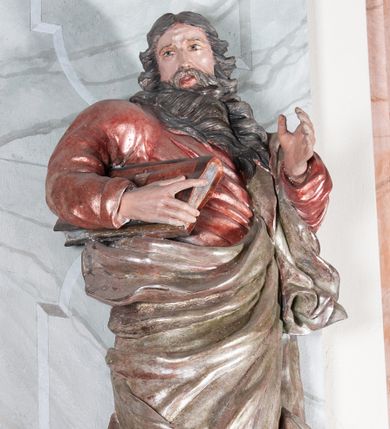 Zdjęcie nr 1: Figura drążona, mocno ścięta z tyłu, przedstawiająca św. Joachima. Święty ustawiony frontalnie, lewa ręka uniesiona i wysunięta przed siebie, a prawą, ugiętą podtrzymuje przy boku księgę. Głowę ma skierowaną w prawą stronę, delikatnie pochyloną. Twarz szeroka z długim nosem o niewielkich oczach patrzących w bok oraz delikatnie rozchylonych ustach; okolona długą brodą w postaci silnie skręconych dwóch pukli opadających diagonalnie na pierś. Włosy długie z przedziałkiem na środku głowy, kręcone, opadają na plecy i lewe ramię. Święty ubrany jest w czerwoną, silnie przylegającą do ciała suknię z wąskimi rękawami oraz brązowy płaszcz przerzucony przez lewe ramię, opadający przez plecy, otaczający postać i odsłaniający klatkę piersiową. Tkanina ekspresyjnie drapowana, obficie fałdowana z rozwianym spodem. Polichromia naturalistyczna w partiach ciała.