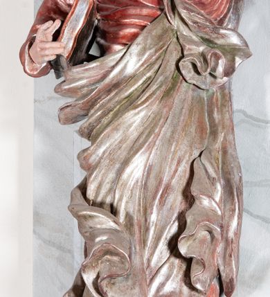 Zdjęcie nr 2: Figura drążona, mocno ścięta z tyłu, przedstawiająca św. Joachima. Święty ustawiony frontalnie, lewa ręka uniesiona i wysunięta przed siebie, a prawą, ugiętą podtrzymuje przy boku księgę. Głowę ma skierowaną w prawą stronę, delikatnie pochyloną. Twarz szeroka z długim nosem o niewielkich oczach patrzących w bok oraz delikatnie rozchylonych ustach; okolona długą brodą w postaci silnie skręconych dwóch pukli opadających diagonalnie na pierś. Włosy długie z przedziałkiem na środku głowy, kręcone, opadają na plecy i lewe ramię. Święty ubrany jest w czerwoną, silnie przylegającą do ciała suknię z wąskimi rękawami oraz brązowy płaszcz przerzucony przez lewe ramię, opadający przez plecy, otaczający postać i odsłaniający klatkę piersiową. Tkanina ekspresyjnie drapowana, obficie fałdowana z rozwianym spodem. Polichromia naturalistyczna w partiach ciała.