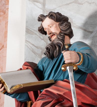 Zdjęcie nr 5: Figura drążona, ścięta z tyłu, przedstawiająca św. Pawła. Święty ustawiony frontalnie, w kontrapoście, z lewą nogą ugiętą w kolanie, o esowato wygiętej sylwetce, z głową zwróconą z prawo. W prawej dłoni trzyma otwartą księgę, a lewą wspiera się na mieczu. Twarz ascetyczna, niewielkie półprzymknięte oczy, delikatnie rozchylone usta, okolona długą brodą opadającą w postaci skręconych pasm. Włosy średniej długości, mocno kręcone, jasnobrązowe. Święty ubrany jest w niebieską suknię, przepasaną w talii, ze złoconą lamówką u spodu oraz czerwony płaszcz podbity złotem, zawinięty na prawym ramieniu, opadający przez plecy na prawą nogę. Tkanina silnie drapowana w głęboko rzeźbione fałdy, ekspresyjnie kształtowana, miejscami przylegająca do ciała. Polichromia ciała naturalistyczna, detale złocone, miecz srebrzony i złocony.