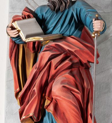 Zdjęcie nr 3: Figura drążona, ścięta z tyłu, przedstawiająca św. Pawła. Święty ustawiony frontalnie, w kontrapoście, z lewą nogą ugiętą w kolanie, o esowato wygiętej sylwetce, z głową zwróconą z prawo. W prawej dłoni trzyma otwartą księgę, a lewą wspiera się na mieczu. Twarz ascetyczna, niewielkie półprzymknięte oczy, delikatnie rozchylone usta, okolona długą brodą opadającą w postaci skręconych pasm. Włosy średniej długości, mocno kręcone, jasnobrązowe. Święty ubrany jest w niebieską suknię, przepasaną w talii, ze złoconą lamówką u spodu oraz czerwony płaszcz podbity złotem, zawinięty na prawym ramieniu, opadający przez plecy na prawą nogę. Tkanina silnie drapowana w głęboko rzeźbione fałdy, ekspresyjnie kształtowana, miejscami przylegająca do ciała. Polichromia ciała naturalistyczna, detale złocone, miecz srebrzony i złocony.