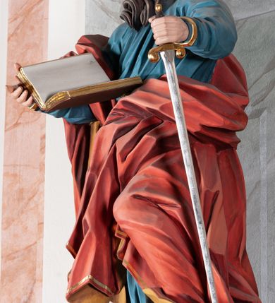 Zdjęcie nr 4: Figura drążona, ścięta z tyłu, przedstawiająca św. Pawła. Święty ustawiony frontalnie, w kontrapoście, z lewą nogą ugiętą w kolanie, o esowato wygiętej sylwetce, z głową zwróconą z prawo. W prawej dłoni trzyma otwartą księgę, a lewą wspiera się na mieczu. Twarz ascetyczna, niewielkie półprzymknięte oczy, delikatnie rozchylone usta, okolona długą brodą opadającą w postaci skręconych pasm. Włosy średniej długości, mocno kręcone, jasnobrązowe. Święty ubrany jest w niebieską suknię, przepasaną w talii, ze złoconą lamówką u spodu oraz czerwony płaszcz podbity złotem, zawinięty na prawym ramieniu, opadający przez plecy na prawą nogę. Tkanina silnie drapowana w głęboko rzeźbione fałdy, ekspresyjnie kształtowana, miejscami przylegająca do ciała. Polichromia ciała naturalistyczna, detale złocone, miecz srebrzony i złocony.