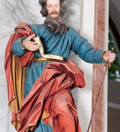 Zdjęcie nr 2: Figura drążona, ścięta z tyłu, przedstawiająca św. Pawła. Święty ustawiony frontalnie, w kontrapoście, z lewą nogą ugiętą w kolanie, o esowato wygiętej sylwetce, z głową zwróconą z prawo. W prawej dłoni trzyma otwartą księgę, a lewą wspiera się na mieczu. Twarz ascetyczna, niewielkie półprzymknięte oczy, delikatnie rozchylone usta, okolona długą brodą opadającą w postaci skręconych pasm. Włosy średniej długości, mocno kręcone, jasnobrązowe. Święty ubrany jest w niebieską suknię, przepasaną w talii, ze złoconą lamówką u spodu oraz czerwony płaszcz podbity złotem, zawinięty na prawym ramieniu, opadający przez plecy na prawą nogę. Tkanina silnie drapowana w głęboko rzeźbione fałdy, ekspresyjnie kształtowana, miejscami przylegająca do ciała. Polichromia ciała naturalistyczna, detale złocone, miecz srebrzony i złocony.