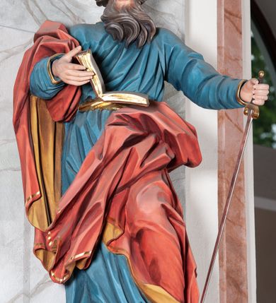 Zdjęcie nr 1: Figura drążona, ścięta z tyłu, przedstawiająca św. Pawła. Święty ustawiony frontalnie, w kontrapoście, z lewą nogą ugiętą w kolanie, o esowato wygiętej sylwetce, z głową zwróconą z prawo. W prawej dłoni trzyma otwartą księgę, a lewą wspiera się na mieczu. Twarz ascetyczna, niewielkie półprzymknięte oczy, delikatnie rozchylone usta, okolona długą brodą opadającą w postaci skręconych pasm. Włosy średniej długości, mocno kręcone, jasnobrązowe. Święty ubrany jest w niebieską suknię, przepasaną w talii, ze złoconą lamówką u spodu oraz czerwony płaszcz podbity złotem, zawinięty na prawym ramieniu, opadający przez plecy na prawą nogę. Tkanina silnie drapowana w głęboko rzeźbione fałdy, ekspresyjnie kształtowana, miejscami przylegająca do ciała. Polichromia ciała naturalistyczna, detale złocone, miecz srebrzony i złocony.