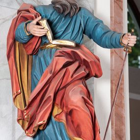 Zdjęcie nr 1: Figura drążona, ścięta z tyłu, przedstawiająca św. Pawła. Święty ustawiony frontalnie, w kontrapoście, z lewą nogą ugiętą w kolanie, o esowato wygiętej sylwetce, z głową zwróconą z prawo. W prawej dłoni trzyma otwartą księgę, a lewą wspiera się na mieczu. Twarz ascetyczna, niewielkie półprzymknięte oczy, delikatnie rozchylone usta, okolona długą brodą opadającą w postaci skręconych pasm. Włosy średniej długości, mocno kręcone, jasnobrązowe. Święty ubrany jest w niebieską suknię, przepasaną w talii, ze złoconą lamówką u spodu oraz czerwony płaszcz podbity złotem, zawinięty na prawym ramieniu, opadający przez plecy na prawą nogę. Tkanina silnie drapowana w głęboko rzeźbione fałdy, ekspresyjnie kształtowana, miejscami przylegająca do ciała. Polichromia ciała naturalistyczna, detale złocone, miecz srebrzony i złocony.
