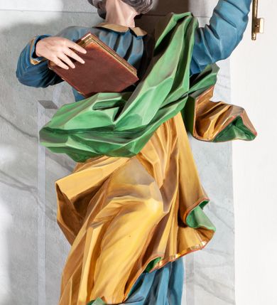 Zdjęcie nr 1: Rzeźba drążona, ścięta z tyłu, przedstawiająca św. Piotra. Święty ustawiony frontalnie, o esowato wygiętej sylwetce, z ugiętą prawą nogą, opartą o stopień. W lewej uniesionej wysoko ręce trzyma dwa klucze, prawą  podtrzymuje opartą o bok księgę. Głowa skierowana trzy czwarte w lewo i uniesiona w górę. Twarz ascetyczna, oczy uniesione w górę, rozchylone usta; okolona gęstą brodą, układającą się w pukle. Włosy siwe w postaci silnie skręconych loków po bokach i na tyle głowy oraz pukiel na czole. Święty ubrany jest w niebieską suknię z kołnierzykiem oraz przerzucony diagonalnie przez lewe ramię żółty płaszcz ze złoconą lamówką i zieloną podszewką. Tkanina ekspresyjnie kształtowana, silnie drapowana, miejscami przylegająca do ciała. Polichromia naturalistyczna, atrybuty i lamówka złocone.