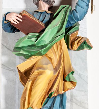 Zdjęcie nr 2: Rzeźba drążona, ścięta z tyłu, przedstawiająca św. Piotra. Święty ustawiony frontalnie, o esowato wygiętej sylwetce, z ugiętą prawą nogą, opartą o stopień. W lewej uniesionej wysoko ręce trzyma dwa klucze, prawą  podtrzymuje opartą o bok księgę. Głowa skierowana trzy czwarte w lewo i uniesiona w górę. Twarz ascetyczna, oczy uniesione w górę, rozchylone usta; okolona gęstą brodą, układającą się w pukle. Włosy siwe w postaci silnie skręconych loków po bokach i na tyle głowy oraz pukiel na czole. Święty ubrany jest w niebieską suknię z kołnierzykiem oraz przerzucony diagonalnie przez lewe ramię żółty płaszcz ze złoconą lamówką i zieloną podszewką. Tkanina ekspresyjnie kształtowana, silnie drapowana, miejscami przylegająca do ciała. Polichromia naturalistyczna, atrybuty i lamówka złocone.