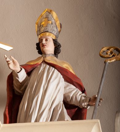 Zdjęcie nr 2: Rzeźba pełnoplastyczna, figura zwrócona trzy czwarte w prawo, ustawiona w kontrapoście z lewą nogą ugiętą w kolanie. W lewej ręce trzyma pastorał, prawą wyciąga przed siebie. Twarz szeroka, z długim i orlim nosem, delikatnie otwartymi ustami, z półprzymkniętymi oczami. Włosy średniej długości, ciemnobrązowe, zawinięte do tyłu w postaci loków. Święty ubrany jest w białą albę oraz złoty płaszcz z czerwoną podszewką spięty na piersi. Na głowie ma założoną infułę ze złoconym krzyżykiem greckim na froncie. U jego stóp, po lewej stronie znajduje się Piotrowin, ukazany w półpostaci, odchylony do tyłu z jedną ręką wzniesioną ku św. Stanisławowi. Polichromia naturalistyczna, detale stroju i atrybutów złocone.