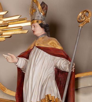 Zdjęcie nr 1: Rzeźba pełnoplastyczna, figura zwrócona trzy czwarte w prawo, ustawiona w kontrapoście z lewą nogą ugiętą w kolanie. W lewej ręce trzyma pastorał, prawą wyciąga przed siebie. Twarz szeroka, z długim i orlim nosem, delikatnie otwartymi ustami, z półprzymkniętymi oczami. Włosy średniej długości, ciemnobrązowe, zawinięte do tyłu w postaci loków. Święty ubrany jest w białą albę oraz złoty płaszcz z czerwoną podszewką spięty na piersi. Na głowie ma założoną infułę ze złoconym krzyżykiem greckim na froncie. U jego stóp, po lewej stronie znajduje się Piotrowin, ukazany w półpostaci, odchylony do tyłu z jedną ręką wzniesioną ku św. Stanisławowi. Polichromia naturalistyczna, detale stroju i atrybutów złocone.