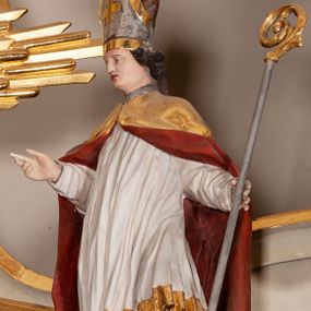 Zdjęcie nr 1: Rzeźba pełnoplastyczna, figura zwrócona trzy czwarte w prawo, ustawiona w kontrapoście z lewą nogą ugiętą w kolanie. W lewej ręce trzyma pastorał, prawą wyciąga przed siebie. Twarz szeroka, z długim i orlim nosem, delikatnie otwartymi ustami, z półprzymkniętymi oczami. Włosy średniej długości, ciemnobrązowe, zawinięte do tyłu w postaci loków. Święty ubrany jest w białą albę oraz złoty płaszcz z czerwoną podszewką spięty na piersi. Na głowie ma założoną infułę ze złoconym krzyżykiem greckim na froncie. U jego stóp, po lewej stronie znajduje się Piotrowin, ukazany w półpostaci, odchylony do tyłu z jedną ręką wzniesioną ku św. Stanisławowi. Polichromia naturalistyczna, detale stroju i atrybutów złocone.