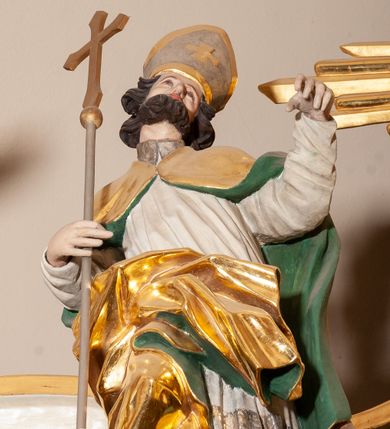 Zdjęcie nr 1: Rzeźba pełnoplastyczna, figura zwrócona trzy czwarte w lewo, ustawiona w pozycji stojącej w silnym kontrapoście, z prawą nogą ugiętą w kolanie, z głową przechyloną w lewą stronę. W prawej ręce trzyma krzyż od długim drzewcu, lewą wyciąga przed siebie. Twarz szeroka, o spiczastym nosie, okolona krótką i ciemnobrązową brodą. Włosy średniej długości, ciemnobrązowe, zasłaniające uszy. Święty ubrany jest w brązową albę, białą komżę oraz złoty płaszcz z zieloną podszewką, spięty na piersi, z silnie drapowaną prawą połą. Na głowie ma srebrzoną infułę ze złoconym krzyżem. Polichromia naturalistyczna, detale stroju złocone.