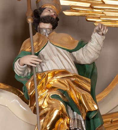Zdjęcie nr 2: Rzeźba pełnoplastyczna, figura zwrócona trzy czwarte w lewo, ustawiona w pozycji stojącej w silnym kontrapoście, z prawą nogą ugiętą w kolanie, z głową przechyloną w lewą stronę. W prawej ręce trzyma krzyż od długim drzewcu, lewą wyciąga przed siebie. Twarz szeroka, o spiczastym nosie, okolona krótką i ciemnobrązową brodą. Włosy średniej długości, ciemnobrązowe, zasłaniające uszy. Święty ubrany jest w brązową albę, białą komżę oraz złoty płaszcz z zieloną podszewką, spięty na piersi, z silnie drapowaną prawą połą. Na głowie ma srebrzoną infułę ze złoconym krzyżem. Polichromia naturalistyczna, detale stroju złocone.