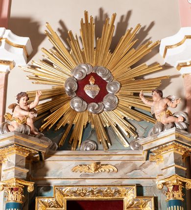 Zdjęcie nr 2: Ołtarz przyścienny, architektoniczny, jednoosiowy, jednokondygnacyjny ze zwieńczeniem. Mensa ołtarzowa skrzyniowa, prostopadłościenna z portatylem w formie kamiennej płytki. Antepedium współczesne, ujęte profilowaną ramą zdobioną żłobionym wałkiem, obite blachą miedzianą, na której odlewana, aplikowana dekoracja przedstawiająca w centrum Baranka Bożego w glorii promienistej z obłokami, po bokach dwie klęczące postacie kobiece, symbole ewangelistów oraz motywy kwiatowe. Tabernakulum w formie prostopadłościennej skrzynki, ustawione na cokole, zamknięte profilowanym gzymsem. Drzwiczki w kształcie stojącego prostokąta z malowanymi czterema scenami z życia Chrystusa. Narożniki obwiedzione złotymi listwami. Nastawa ołtarzowa umieszczona na podwójnym cokole, flankowana dwiema kolumnami, kanelowanymi od jednej czwartej wysokości o kapitelach kompozytowych, dźwigających pełne, wyłamane belkowanie oraz imposty ustawione pod kątem, na których znajduje się para puttów. Po zewnętrznej stronie kolumn cokoły z figurami św. Józefa z Dzieciątkiem po lewej i św. Anny z Marią po prawej. Pole środkowe w formie niszy w kształcie stojącego prostokąta o wciętych górnych narożach z pełnoplastyczną figurą Najświętszego Serca Jezusa. Zwieńczenie w formie promienistej glorii, z otoczonym obłokami medalionem w środku z gorejącym sercem. Cokoły figur i kolumn dekorowane płycinami z esowato wygiętym ornamentem roślinnym. Gzyms impostów zdobiony ornamentem roślinnym, w partii fryzu rzeźbiona uskrzydlona główka anielska. Rama w polu środkowym dekorowana płaskorzeźbionym ornamentem roślinnym. Struktura marmoryzowana w odcieniu różowo-błękitnym, detale architektoniczne polichromowane na niebiesko i czerwono, ornamentyka i profilowania pozłacane i posrebrzane.