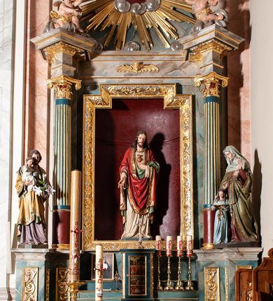 Zdjęcie nr 2: Rzeźba pełna, ustawiona frontalnie na niskim, zielonym postumencie. Chrystus stoi w delikatnym kontrapoście, lewą ręką wskazuje na gorejące na piersi serce, prawą opuszczoną w dół, ukazuje ranę na dłoni. Ma podłużną twarz, z delikatnie przymkniętymi oczami, długim i wąskim nosem, okoloną krótką brodą zakończoną w dwa pukle; włosy długie, z przedziałkiem  na środku głowy, opadają na ramiona w postaci loków. Chrystus ubrany jest w długą, sięgającą stóp, białą i silnie drapowaną suknię, o szerokich rękawach, ze złotą lamówką pod szyją. Na ramiona ma narzucony czerwony płaszcz, podtrzymywany lewą ręką, z dekorowaną i złoconą bordiurą oraz zielonym podbiciem. Polichromia naturalistyczna w odsłoniętych partiach ciała, detal stroju złocony.