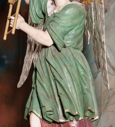 Zdjęcie nr 4: Rzeźba drążona, przedstawiająca anioła z arma Christi w dłoniach. Figura ustawiona frontalnie z prawą nogą wysuniętą do przodu, z głową skierowaną w lewą stronę. W uniesionych po swojej prawej stronie dłoniach trzyma drabinę i koronę cierniową. Twarz szeroka z wysokim czołem, prostym nosem, wzrokiem skierowanym w dół i delikatnie rozchylonymi ustami. Włosy długie, brązowe z przedziałkiem pośrodku, opadające na ramiona. Anioł ubrany jest w czerwoną, długą szatę spodnią, silnie drapowaną, odsłaniającą prawą nogę, na niej ma założoną zieloną tunikę z białym kołnierzykiem, a na stopach pomarańczowe, rzymskie sandały. Polichromia naturalistyczna, skrzydła posrebrzane i pozłacane, atrybuty złocone.

