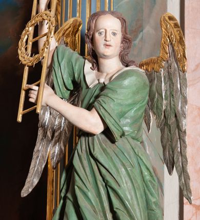 Zdjęcie nr 1: Rzeźba drążona, przedstawiająca anioła z arma Christi w dłoniach. Figura ustawiona frontalnie z prawą nogą wysuniętą do przodu, z głową skierowaną w lewą stronę. W uniesionych po swojej prawej stronie dłoniach trzyma drabinę i koronę cierniową. Twarz szeroka z wysokim czołem, prostym nosem, wzrokiem skierowanym w dół i delikatnie rozchylonymi ustami. Włosy długie, brązowe z przedziałkiem pośrodku, opadające na ramiona. Anioł ubrany jest w czerwoną, długą szatę spodnią, silnie drapowaną, odsłaniającą prawą nogę, na niej ma założoną zieloną tunikę z białym kołnierzykiem, a na stopach pomarańczowe, rzymskie sandały. Polichromia naturalistyczna, skrzydła posrebrzane i pozłacane, atrybuty złocone.

