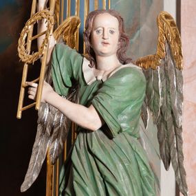 Zdjęcie nr 1: Rzeźba drążona, przedstawiająca anioła z arma Christi w dłoniach. Figura ustawiona frontalnie z prawą nogą wysuniętą do przodu, z głową skierowaną w lewą stronę. W uniesionych po swojej prawej stronie dłoniach trzyma drabinę i koronę cierniową. Twarz szeroka z wysokim czołem, prostym nosem, wzrokiem skierowanym w dół i delikatnie rozchylonymi ustami. Włosy długie, brązowe z przedziałkiem pośrodku, opadające na ramiona. Anioł ubrany jest w czerwoną, długą szatę spodnią, silnie drapowaną, odsłaniającą prawą nogę, na niej ma założoną zieloną tunikę z białym kołnierzykiem, a na stopach pomarańczowe, rzymskie sandały. Polichromia naturalistyczna, skrzydła posrebrzane i pozłacane, atrybuty złocone.

