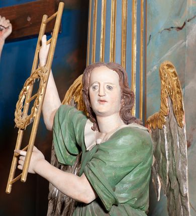 Zdjęcie nr 6: Rzeźba drążona, przedstawiająca anioła z arma Christi w dłoniach. Figura ustawiona frontalnie z prawą nogą wysuniętą do przodu, z głową skierowaną w lewą stronę. W uniesionych po swojej prawej stronie dłoniach trzyma drabinę i koronę cierniową. Twarz szeroka z wysokim czołem, prostym nosem, wzrokiem skierowanym w dół i delikatnie rozchylonymi ustami. Włosy długie, brązowe z przedziałkiem pośrodku, opadające na ramiona. Anioł ubrany jest w czerwoną, długą szatę spodnią, silnie drapowaną, odsłaniającą prawą nogę, na niej ma założoną zieloną tunikę z białym kołnierzykiem, a na stopach pomarańczowe, rzymskie sandały. Polichromia naturalistyczna, skrzydła posrebrzane i pozłacane, atrybuty złocone.

