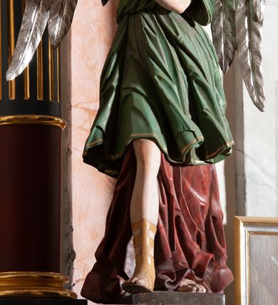 Zdjęcie nr 5: Rzeźba drążona, przedstawiająca anioła z arma Christi w dłoniach. Figura ustawiona frontalnie z prawą nogą wysuniętą do przodu, z głową skierowaną w lewą stronę. W uniesionych po swojej prawej stronie dłoniach trzyma drabinę i koronę cierniową. Twarz szeroka z wysokim czołem, prostym nosem, wzrokiem skierowanym w dół i delikatnie rozchylonymi ustami. Włosy długie, brązowe z przedziałkiem pośrodku, opadające na ramiona. Anioł ubrany jest w czerwoną, długą szatę spodnią, silnie drapowaną, odsłaniającą prawą nogę, na niej ma założoną zieloną tunikę z białym kołnierzykiem, a na stopach pomarańczowe, rzymskie sandały. Polichromia naturalistyczna, skrzydła posrebrzane i pozłacane, atrybuty złocone.

