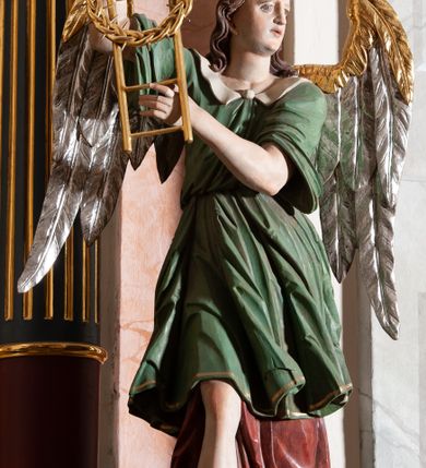 Zdjęcie nr 3: Rzeźba drążona, przedstawiająca anioła z arma Christi w dłoniach. Figura ustawiona frontalnie z prawą nogą wysuniętą do przodu, z głową skierowaną w lewą stronę. W uniesionych po swojej prawej stronie dłoniach trzyma drabinę i koronę cierniową. Twarz szeroka z wysokim czołem, prostym nosem, wzrokiem skierowanym w dół i delikatnie rozchylonymi ustami. Włosy długie, brązowe z przedziałkiem pośrodku, opadające na ramiona. Anioł ubrany jest w czerwoną, długą szatę spodnią, silnie drapowaną, odsłaniającą prawą nogę, na niej ma założoną zieloną tunikę z białym kołnierzykiem, a na stopach pomarańczowe, rzymskie sandały. Polichromia naturalistyczna, skrzydła posrebrzane i pozłacane, atrybuty złocone.

