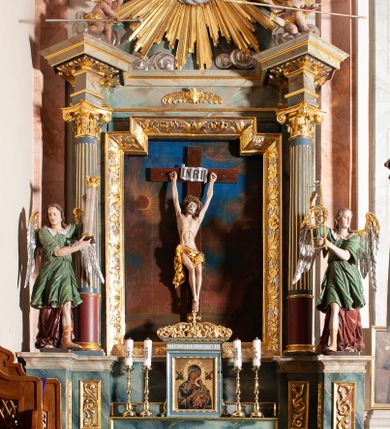 Zdjęcie nr 8: Rzeźba drążona, przedstawiająca anioła z arma Christi w dłoniach. Figura ustawiona frontalnie z prawą nogą wysuniętą do przodu, z głową skierowaną w lewą stronę. W uniesionych po swojej prawej stronie dłoniach trzyma drabinę i koronę cierniową. Twarz szeroka z wysokim czołem, prostym nosem, wzrokiem skierowanym w dół i delikatnie rozchylonymi ustami. Włosy długie, brązowe z przedziałkiem pośrodku, opadające na ramiona. Anioł ubrany jest w czerwoną, długą szatę spodnią, silnie drapowaną, odsłaniającą prawą nogę, na niej ma założoną zieloną tunikę z białym kołnierzykiem, a na stopach pomarańczowe, rzymskie sandały. Polichromia naturalistyczna, skrzydła posrebrzane i pozłacane, atrybuty złocone.

