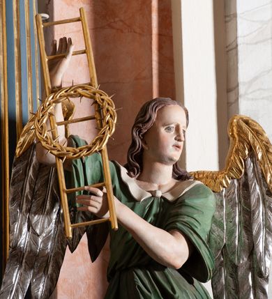 Zdjęcie nr 7: Rzeźba drążona, przedstawiająca anioła z arma Christi w dłoniach. Figura ustawiona frontalnie z prawą nogą wysuniętą do przodu, z głową skierowaną w lewą stronę. W uniesionych po swojej prawej stronie dłoniach trzyma drabinę i koronę cierniową. Twarz szeroka z wysokim czołem, prostym nosem, wzrokiem skierowanym w dół i delikatnie rozchylonymi ustami. Włosy długie, brązowe z przedziałkiem pośrodku, opadające na ramiona. Anioł ubrany jest w czerwoną, długą szatę spodnią, silnie drapowaną, odsłaniającą prawą nogę, na niej ma założoną zieloną tunikę z białym kołnierzykiem, a na stopach pomarańczowe, rzymskie sandały. Polichromia naturalistyczna, skrzydła posrebrzane i pozłacane, atrybuty złocone.

