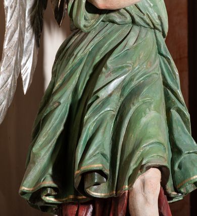 Zdjęcie nr 6: Rzeźba pełnoplastyczna, drążona, przedstawiająca anioła z arma Christi w dłoniach. Figura ustawiona frontalnie z lewą nogą wysuniętą do przodu, z głową skierowaną w prawą stronę. W uniesionych po swojej lewej stronie dłoniach trzyma kolumnę biczowania. Twarz szeroka z wysokim czołem, prostym nosem, wzrokiem skierowanym w dół i delikatnie rozchylonymi ustami. Włosy długie, ciemnobrązowe z przedziałkiem pośrodku, opadające na ramiona.  Anioł jest ubrany w czerwoną, długą szatę spodnią, silnie drapowaną, na niej ma założoną zieloną tunikę z białym kołnierzykiem, na stopach pomarańczowe, rzymskie sandały. Kolumna ustawiona na bazie, trzon gładki, kapitel koryncki. Polichromia naturalistyczna, skrzydła posrebrzane i pozłacane.
