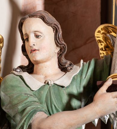 Zdjęcie nr 5: Rzeźba pełnoplastyczna, drążona, przedstawiająca anioła z arma Christi w dłoniach. Figura ustawiona frontalnie z lewą nogą wysuniętą do przodu, z głową skierowaną w prawą stronę. W uniesionych po swojej lewej stronie dłoniach trzyma kolumnę biczowania. Twarz szeroka z wysokim czołem, prostym nosem, wzrokiem skierowanym w dół i delikatnie rozchylonymi ustami. Włosy długie, ciemnobrązowe z przedziałkiem pośrodku, opadające na ramiona.  Anioł jest ubrany w czerwoną, długą szatę spodnią, silnie drapowaną, na niej ma założoną zieloną tunikę z białym kołnierzykiem, na stopach pomarańczowe, rzymskie sandały. Kolumna ustawiona na bazie, trzon gładki, kapitel koryncki. Polichromia naturalistyczna, skrzydła posrebrzane i pozłacane.