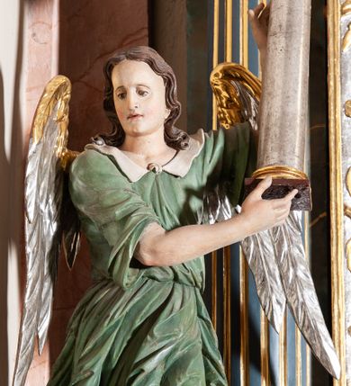 Zdjęcie nr 3: Rzeźba pełnoplastyczna, drążona, przedstawiająca anioła z arma Christi w dłoniach. Figura ustawiona frontalnie z lewą nogą wysuniętą do przodu, z głową skierowaną w prawą stronę. W uniesionych po swojej lewej stronie dłoniach trzyma kolumnę biczowania. Twarz szeroka z wysokim czołem, prostym nosem, wzrokiem skierowanym w dół i delikatnie rozchylonymi ustami. Włosy długie, ciemnobrązowe z przedziałkiem pośrodku, opadające na ramiona.  Anioł jest ubrany w czerwoną, długą szatę spodnią, silnie drapowaną, na niej ma założoną zieloną tunikę z białym kołnierzykiem, na stopach pomarańczowe, rzymskie sandały. Kolumna ustawiona na bazie, trzon gładki, kapitel koryncki. Polichromia naturalistyczna, skrzydła posrebrzane i pozłacane.