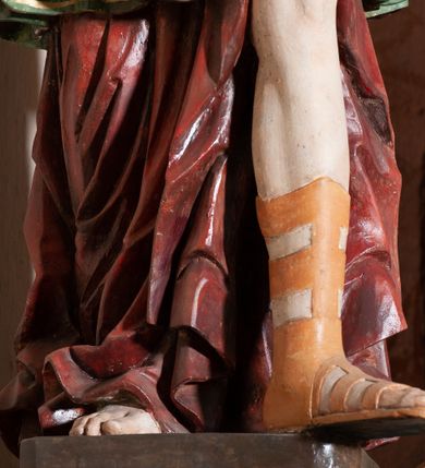 Zdjęcie nr 7: Rzeźba pełnoplastyczna, drążona, przedstawiająca anioła z arma Christi w dłoniach. Figura ustawiona frontalnie z lewą nogą wysuniętą do przodu, z głową skierowaną w prawą stronę. W uniesionych po swojej lewej stronie dłoniach trzyma kolumnę biczowania. Twarz szeroka z wysokim czołem, prostym nosem, wzrokiem skierowanym w dół i delikatnie rozchylonymi ustami. Włosy długie, ciemnobrązowe z przedziałkiem pośrodku, opadające na ramiona.  Anioł jest ubrany w czerwoną, długą szatę spodnią, silnie drapowaną, na niej ma założoną zieloną tunikę z białym kołnierzykiem, na stopach pomarańczowe, rzymskie sandały. Kolumna ustawiona na bazie, trzon gładki, kapitel koryncki. Polichromia naturalistyczna, skrzydła posrebrzane i pozłacane.