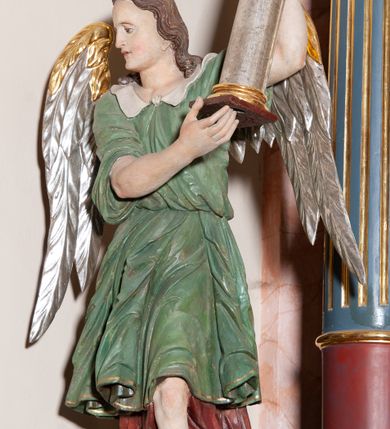 Zdjęcie nr 2: Rzeźba pełnoplastyczna, drążona, przedstawiająca anioła z arma Christi w dłoniach. Figura ustawiona frontalnie z lewą nogą wysuniętą do przodu, z głową skierowaną w prawą stronę. W uniesionych po swojej lewej stronie dłoniach trzyma kolumnę biczowania. Twarz szeroka z wysokim czołem, prostym nosem, wzrokiem skierowanym w dół i delikatnie rozchylonymi ustami. Włosy długie, ciemnobrązowe z przedziałkiem pośrodku, opadające na ramiona.  Anioł jest ubrany w czerwoną, długą szatę spodnią, silnie drapowaną, na niej ma założoną zieloną tunikę z białym kołnierzykiem, na stopach pomarańczowe, rzymskie sandały. Kolumna ustawiona na bazie, trzon gładki, kapitel koryncki. Polichromia naturalistyczna, skrzydła posrebrzane i pozłacane.