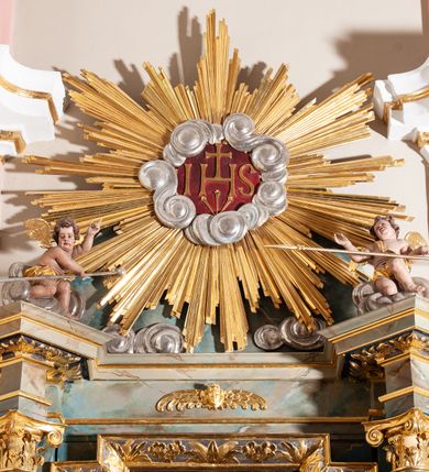 Zdjęcie nr 3: Ołtarz przyścienny, architektoniczny, jednoosiowy, jednokondygnacyjny, ze zwieńczeniem. Mensa ołtarzowa skrzyniowa, prostopadłościenna, z portatylem w formie kamiennej płyty. Antepedium współczesne, ujęte profilowaną ramą zdobioną żłobionym wałkiem, obite blachą miedzianą, na której odlewana, aplikowana dekoracja przedstawiająca w centrum Baranka Bożego w glorii promienistej z obłokami, po bokach dwie klęczące postacie kobiece, symbole ewangelistów oraz motywy kwiatowe. Nastawa ołtarzowa na podwójnym cokole, flankowana dwiema kanelowanymi od jednej czwartej wysokości kolumnami o kapitelach kompozytowych, dźwigających pełne i wyłamane belkowanie oraz imposty ustawione pod kątem, na których znajduje się para puttów z arma Christi. Po zewnętrznej stronie kolumn cokoły z figurami aniołów. Pole środkowe w formie niszy w kształcie stojącego prostokąta o wciętych górnych narożach, gdzie na tle malowanego pejzażu znajduje się rzeźbiony krucyfiks. Zwieńczenie w formie promienistej glorii z otoczonym obłokami medalionem, w którego polu monogram IHS. Cokoły figur i kolumn dekorowane płycinami z esowato wygiętym ornamentem roślinnym. Gzyms impostów zdobiony ornamentem roślinnym, w partii fryzu rzeźbiona uskrzydlona główka anielska. Rama w polu środkowym dekorowana płaskorzeźbionym ornamentem roślinnym. Struktura marmoryzowana w odcieniu różowo-błękitnym, detale architektoniczne polichromowane na niebiesko i czerwono, ornamentyka i profilowania pozłacane i posrebrzane.