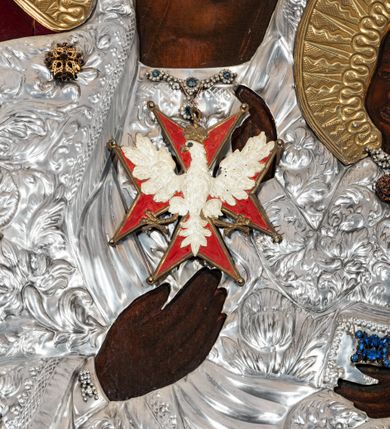 Zdjęcie nr 4: Obraz w kształcie stojącego prostokąta z przedstawieniem Matki Boskiej z Dzieciątkiem w typie Częstochowskiej, w srebrnej sukience odsłaniającej jedynie twarze i dłonie, otoczony szeroką bordiurą z repusowaną dekoracją floralną. Matka Boska ukazana frontalnie z Dzieciątkiem na lewym ręku, prawą rękę ma złożoną na piersi. Twarze o ciemnych karnacjach. Matka Boska delikatnie zwraca głowę w lewo, ma wzrok skierowany w dal, długi i wąski nos, na prawym policzku dwie blizny. Dzieciątko ukazane w pozycji siedzącej, z prawą rączką wzniesioną w geście błogosławieństwa, w lewej trzyma na kolanach zamkniętą księgę. Sukienki drewniane, płaszcz Matki Boskiej bogato dekorowany roślinną dekoracją. Wokół głów obu postaci złote, koliste nimby promieniste z nałożonymi, bardzo ozdobnymi, również złotymi koronami utworzonymi z ornamentu roślinnego dekorowanego kameryzacją. Sukienki obu postaci srebrne, również zdobione ornamentem roślinnym i kameryzacją, na dekolcie Marii aplikowany order Orła Białego. Tło obrazu stanowi ciemnoczerwona tkanina. Przedstawienie obwiedzione szerokim, metalowym pasem srebrnej dekoracji floralnej ze złoconymi plakietami w narożach i połowie wysokości każdego boku ramy. U góry zawieszono rząd orderów, po bokach serca wotywne, na dolnej krawędzi liczne sznury korali. 