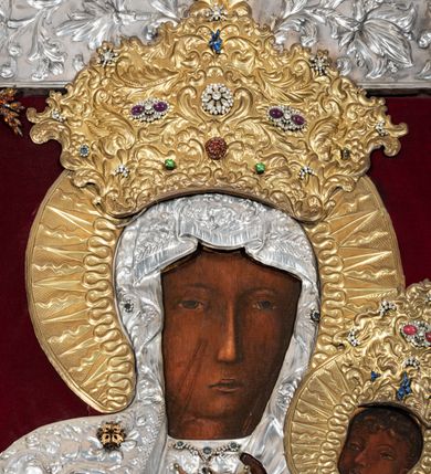 Zdjęcie nr 6: Obraz w kształcie stojącego prostokąta z przedstawieniem Matki Boskiej z Dzieciątkiem w typie Częstochowskiej, w srebrnej sukience odsłaniającej jedynie twarze i dłonie, otoczony szeroką bordiurą z repusowaną dekoracją floralną. Matka Boska ukazana frontalnie z Dzieciątkiem na lewym ręku, prawą rękę ma złożoną na piersi. Twarze o ciemnych karnacjach. Matka Boska delikatnie zwraca głowę w lewo, ma wzrok skierowany w dal, długi i wąski nos, na prawym policzku dwie blizny. Dzieciątko ukazane w pozycji siedzącej, z prawą rączką wzniesioną w geście błogosławieństwa, w lewej trzyma na kolanach zamkniętą księgę. Sukienki drewniane, płaszcz Matki Boskiej bogato dekorowany roślinną dekoracją. Wokół głów obu postaci złote, koliste nimby promieniste z nałożonymi, bardzo ozdobnymi, również złotymi koronami utworzonymi z ornamentu roślinnego dekorowanego kameryzacją. Sukienki obu postaci srebrne, również zdobione ornamentem roślinnym i kameryzacją, na dekolcie Marii aplikowany order Orła Białego. Tło obrazu stanowi ciemnoczerwona tkanina. Przedstawienie obwiedzione szerokim, metalowym pasem srebrnej dekoracji floralnej ze złoconymi plakietami w narożach i połowie wysokości każdego boku ramy. U góry zawieszono rząd orderów, po bokach serca wotywne, na dolnej krawędzi liczne sznury korali. 