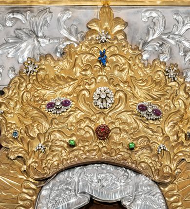 Zdjęcie nr 7: Obraz w kształcie stojącego prostokąta z przedstawieniem Matki Boskiej z Dzieciątkiem w typie Częstochowskiej, w srebrnej sukience odsłaniającej jedynie twarze i dłonie, otoczony szeroką bordiurą z repusowaną dekoracją floralną. Matka Boska ukazana frontalnie z Dzieciątkiem na lewym ręku, prawą rękę ma złożoną na piersi. Twarze o ciemnych karnacjach. Matka Boska delikatnie zwraca głowę w lewo, ma wzrok skierowany w dal, długi i wąski nos, na prawym policzku dwie blizny. Dzieciątko ukazane w pozycji siedzącej, z prawą rączką wzniesioną w geście błogosławieństwa, w lewej trzyma na kolanach zamkniętą księgę. Sukienki drewniane, płaszcz Matki Boskiej bogato dekorowany roślinną dekoracją. Wokół głów obu postaci złote, koliste nimby promieniste z nałożonymi, bardzo ozdobnymi, również złotymi koronami utworzonymi z ornamentu roślinnego dekorowanego kameryzacją. Sukienki obu postaci srebrne, również zdobione ornamentem roślinnym i kameryzacją, na dekolcie Marii aplikowany order Orła Białego. Tło obrazu stanowi ciemnoczerwona tkanina. Przedstawienie obwiedzione szerokim, metalowym pasem srebrnej dekoracji floralnej ze złoconymi plakietami w narożach i połowie wysokości każdego boku ramy. U góry zawieszono rząd orderów, po bokach serca wotywne, na dolnej krawędzi liczne sznury korali. 