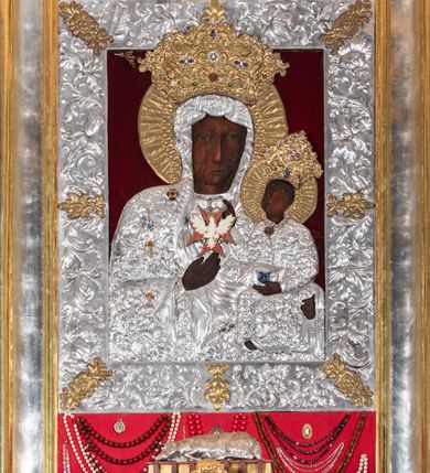 Zdjęcie nr 2: Obraz w kształcie stojącego prostokąta z przedstawieniem Matki Boskiej z Dzieciątkiem w typie Częstochowskiej, w srebrnej sukience odsłaniającej jedynie twarze i dłonie, otoczony szeroką bordiurą z repusowaną dekoracją floralną. Matka Boska ukazana frontalnie z Dzieciątkiem na lewym ręku, prawą rękę ma złożoną na piersi. Twarze o ciemnych karnacjach. Matka Boska delikatnie zwraca głowę w lewo, ma wzrok skierowany w dal, długi i wąski nos, na prawym policzku dwie blizny. Dzieciątko ukazane w pozycji siedzącej, z prawą rączką wzniesioną w geście błogosławieństwa, w lewej trzyma na kolanach zamkniętą księgę. Sukienki drewniane, płaszcz Matki Boskiej bogato dekorowany roślinną dekoracją. Wokół głów obu postaci złote, koliste nimby promieniste z nałożonymi, bardzo ozdobnymi, również złotymi koronami utworzonymi z ornamentu roślinnego dekorowanego kameryzacją. Sukienki obu postaci srebrne, również zdobione ornamentem roślinnym i kameryzacją, na dekolcie Marii aplikowany order Orła Białego. Tło obrazu stanowi ciemnoczerwona tkanina. Przedstawienie obwiedzione szerokim, metalowym pasem srebrnej dekoracji floralnej ze złoconymi plakietami w narożach i połowie wysokości każdego boku ramy. U góry zawieszono rząd orderów, po bokach serca wotywne, na dolnej krawędzi liczne sznury korali. 