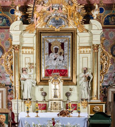 Zdjęcie nr 9: Obraz w kształcie stojącego prostokąta z przedstawieniem Matki Boskiej z Dzieciątkiem w typie Częstochowskiej, w srebrnej sukience odsłaniającej jedynie twarze i dłonie, otoczony szeroką bordiurą z repusowaną dekoracją floralną. Matka Boska ukazana frontalnie z Dzieciątkiem na lewym ręku, prawą rękę ma złożoną na piersi. Twarze o ciemnych karnacjach. Matka Boska delikatnie zwraca głowę w lewo, ma wzrok skierowany w dal, długi i wąski nos, na prawym policzku dwie blizny. Dzieciątko ukazane w pozycji siedzącej, z prawą rączką wzniesioną w geście błogosławieństwa, w lewej trzyma na kolanach zamkniętą księgę. Sukienki drewniane, płaszcz Matki Boskiej bogato dekorowany roślinną dekoracją. Wokół głów obu postaci złote, koliste nimby promieniste z nałożonymi, bardzo ozdobnymi, również złotymi koronami utworzonymi z ornamentu roślinnego dekorowanego kameryzacją. Sukienki obu postaci srebrne, również zdobione ornamentem roślinnym i kameryzacją, na dekolcie Marii aplikowany order Orła Białego. Tło obrazu stanowi ciemnoczerwona tkanina. Przedstawienie obwiedzione szerokim, metalowym pasem srebrnej dekoracji floralnej ze złoconymi plakietami w narożach i połowie wysokości każdego boku ramy. U góry zawieszono rząd orderów, po bokach serca wotywne, na dolnej krawędzi liczne sznury korali. 