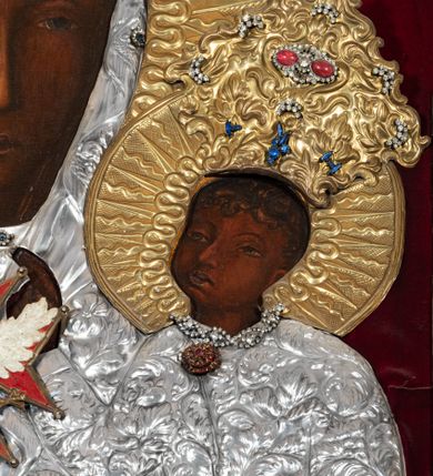 Zdjęcie nr 8: Obraz w kształcie stojącego prostokąta z przedstawieniem Matki Boskiej z Dzieciątkiem w typie Częstochowskiej, w srebrnej sukience odsłaniającej jedynie twarze i dłonie, otoczony szeroką bordiurą z repusowaną dekoracją floralną. Matka Boska ukazana frontalnie z Dzieciątkiem na lewym ręku, prawą rękę ma złożoną na piersi. Twarze o ciemnych karnacjach. Matka Boska delikatnie zwraca głowę w lewo, ma wzrok skierowany w dal, długi i wąski nos, na prawym policzku dwie blizny. Dzieciątko ukazane w pozycji siedzącej, z prawą rączką wzniesioną w geście błogosławieństwa, w lewej trzyma na kolanach zamkniętą księgę. Sukienki drewniane, płaszcz Matki Boskiej bogato dekorowany roślinną dekoracją. Wokół głów obu postaci złote, koliste nimby promieniste z nałożonymi, bardzo ozdobnymi, również złotymi koronami utworzonymi z ornamentu roślinnego dekorowanego kameryzacją. Sukienki obu postaci srebrne, również zdobione ornamentem roślinnym i kameryzacją, na dekolcie Marii aplikowany order Orła Białego. Tło obrazu stanowi ciemnoczerwona tkanina. Przedstawienie obwiedzione szerokim, metalowym pasem srebrnej dekoracji floralnej ze złoconymi plakietami w narożach i połowie wysokości każdego boku ramy. U góry zawieszono rząd orderów, po bokach serca wotywne, na dolnej krawędzi liczne sznury korali. 