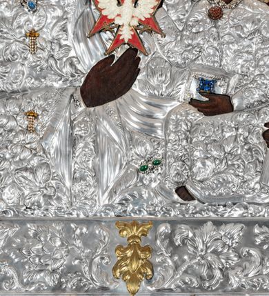 Zdjęcie nr 5: Obraz w kształcie stojącego prostokąta z przedstawieniem Matki Boskiej z Dzieciątkiem w typie Częstochowskiej, w srebrnej sukience odsłaniającej jedynie twarze i dłonie, otoczony szeroką bordiurą z repusowaną dekoracją floralną. Matka Boska ukazana frontalnie z Dzieciątkiem na lewym ręku, prawą rękę ma złożoną na piersi. Twarze o ciemnych karnacjach. Matka Boska delikatnie zwraca głowę w lewo, ma wzrok skierowany w dal, długi i wąski nos, na prawym policzku dwie blizny. Dzieciątko ukazane w pozycji siedzącej, z prawą rączką wzniesioną w geście błogosławieństwa, w lewej trzyma na kolanach zamkniętą księgę. Sukienki drewniane, płaszcz Matki Boskiej bogato dekorowany roślinną dekoracją. Wokół głów obu postaci złote, koliste nimby promieniste z nałożonymi, bardzo ozdobnymi, również złotymi koronami utworzonymi z ornamentu roślinnego dekorowanego kameryzacją. Sukienki obu postaci srebrne, również zdobione ornamentem roślinnym i kameryzacją, na dekolcie Marii aplikowany order Orła Białego. Tło obrazu stanowi ciemnoczerwona tkanina. Przedstawienie obwiedzione szerokim, metalowym pasem srebrnej dekoracji floralnej ze złoconymi plakietami w narożach i połowie wysokości każdego boku ramy. U góry zawieszono rząd orderów, po bokach serca wotywne, na dolnej krawędzi liczne sznury korali. 