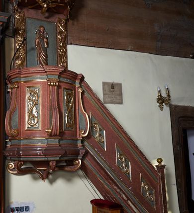 Zdjęcie nr 2: Ambona zawieszona na północnej ścianie prezbiterium. Kosz na rzucie owalu, z wydzielonym profilowanym cokołem i gzymsem, opięty wolutami umieszczonymi na tle pilastrów; wsparty na nadwieszonej podstawie o formie odwróconego spłaszczonego stożka, ujętego pięcioma wolutowo wygiętymi kabłąkami. Zaplecek prostokątny, łączący się z owalnym baldachimem zwieńczonym krzyżem ustawionym na czterech kabłąkach. Baldachim od dołu dekorowany lambrekinem. Od strony wschodniej schody z balustradą zdobioną romboidalnymi płycinami z aplikowanym ornamentem roślinnym. Płyciny kosza oraz boki zaplecka dekorowane wicią akantu. Pośrodku zaplecka płaskorzeźbiona figura Chrystusa Dobrego Pasterza, w podniebiu rzeźbiona Gołębica Ducha Świętego otoczona promieniami. Całość struktury ambony polichromowana na kolor ciemnobrązowy oraz ciemnozielony w partii płycin, detal i profilowania pozłacane.